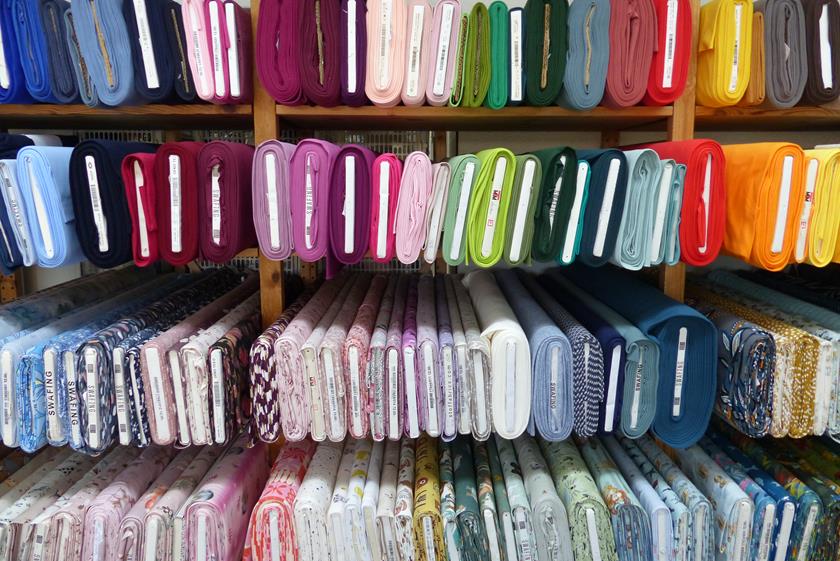 ine grosse Auswahl an verschiedenen Stoffen mit bunten Farben und Mustern, ordentlich gefaltet und sortiert im Verkaufsregal des Lanavera Ladens.
